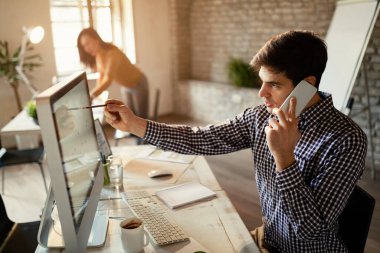 Genç işadamı ofisteki cep telefonuyla iletişim kurarken bilgisayardaki çizelgeleri analiz ediyor..