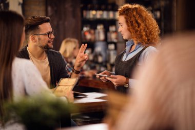 Young man talking to a waitress and making an order while being with girlfriend in a bar. 