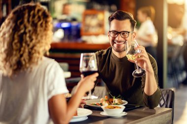 Young happy man drinking wine and talking to his girlfriend while having lunch in a bar. 