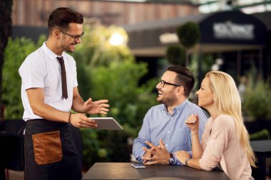 Happy couple talking to a waiter while he's showing them menu on digital tablet in a cafe. 