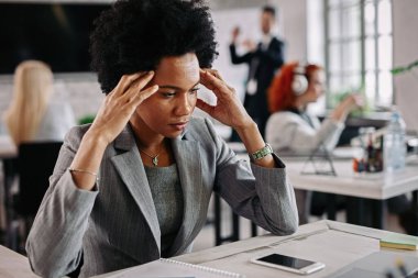 Pensive black businesswoman holding her head in pain while having some problems at work. There are people in the background. 