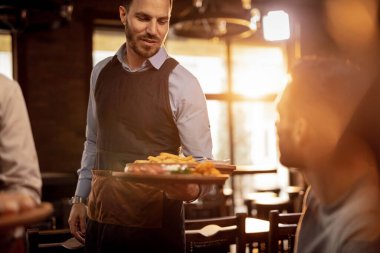 Young waiter serving customers in a restaurant and bringing food at their table. 