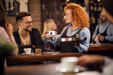 Smiling redhead waitress serving customers and brining coffee to their table in a cafe.