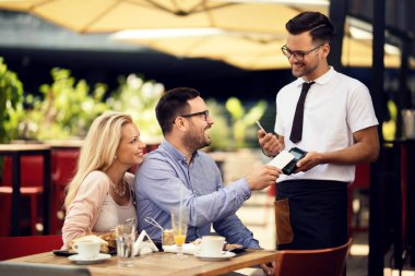 Happy man using smart phone and paying a bill to a waiter while being with his girlfriend in a restaurant. 