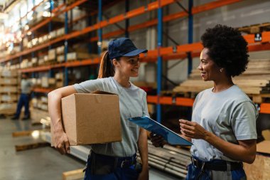 Happy female warehouse workers talking to each other while preparing shipment for distribution in industrial warehouse. 