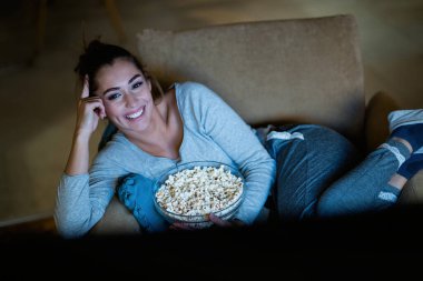 Young happy woman watching TV while relaxing in armchair at night at home. 