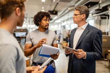 Mid adult engineer using touchpad and communicating with African American worker about product reports in industrial building. 