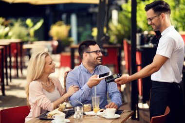 Happy waiter taking a smart phone payment from his guests in a restaurant. 