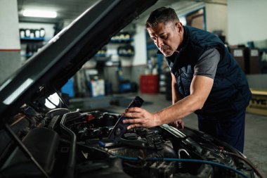 Car mechanic using lamp while examining engine under the hood at auto repair shop. 