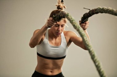 Dedicated female athlete having cross training in gym and exercising with battle rope.