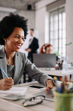 Smiling black businesswoman working on a computer and taking notes at her office desk. There are people in the background. 