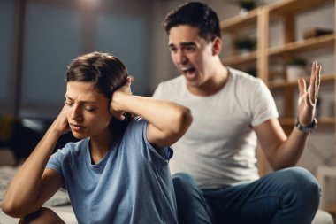 Young man arguing with his girlfriend and screaming at her while she is covering her ears. Focus is on woman. 