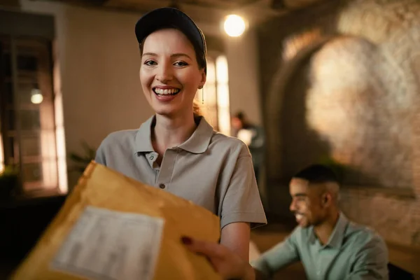 Happy female courier delivering package and looking at camera. There are people in the background.