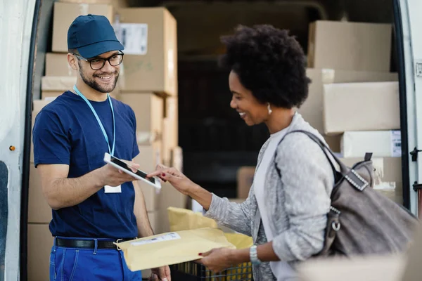 Happy delivery man giving digital tablet to African American woman to sing for a delivery. 