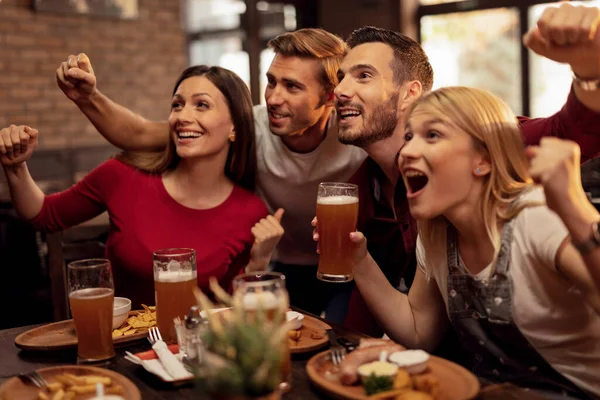 Excited young people watching sports match on TV while drinking beer and eating in a pub.