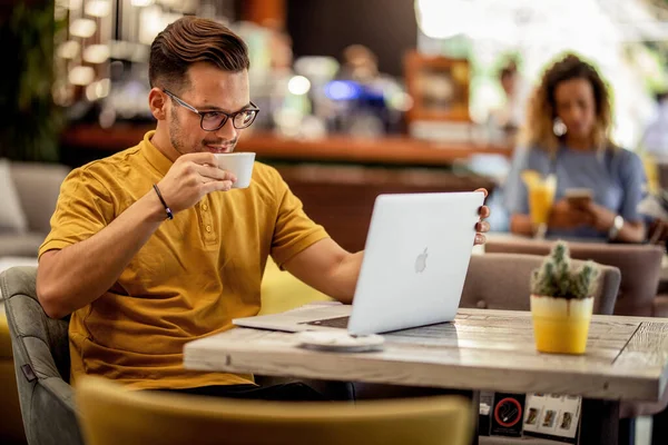 Young freelance worker drinking coffee while working on laptop in a cafe.