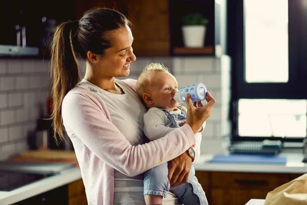 Young caring mother using baby bottle and feeding her baby son in the kitchen. 