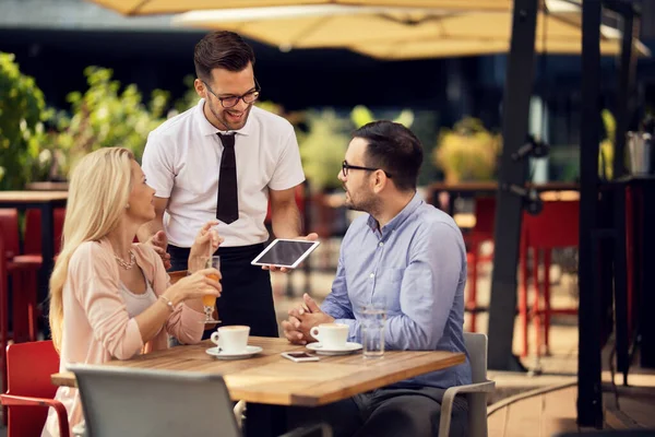 Young happy waiter communicating with guests in an outdoor cafe.