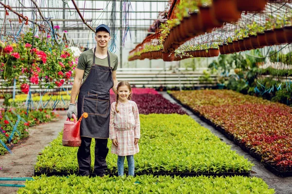 Full length of young happy worker and small girl standing at plant nursery and looking at camera.