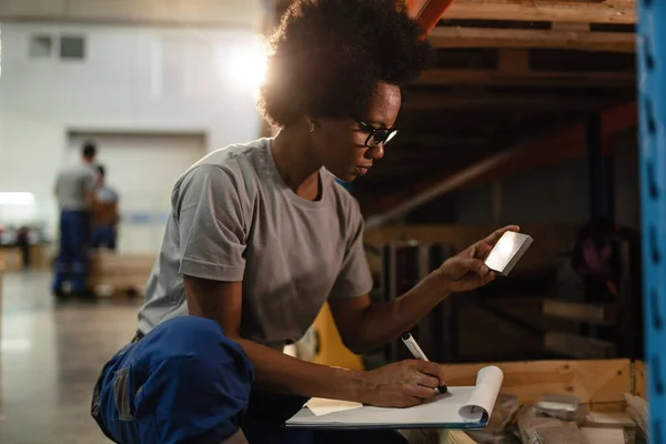 African American warehouse worker making a record of products in shipment list in industrial storage compartment.