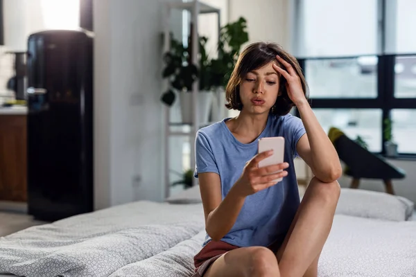 Young woman frowning while typing text message on mobile phone in the bedroom.