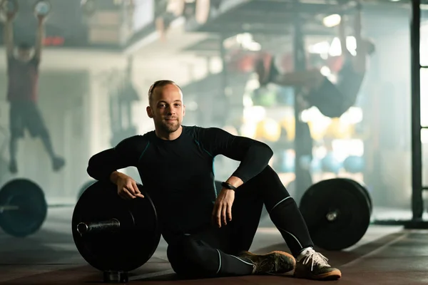 Smiling male athlete relaxing on the floor while having strength training in a gym.