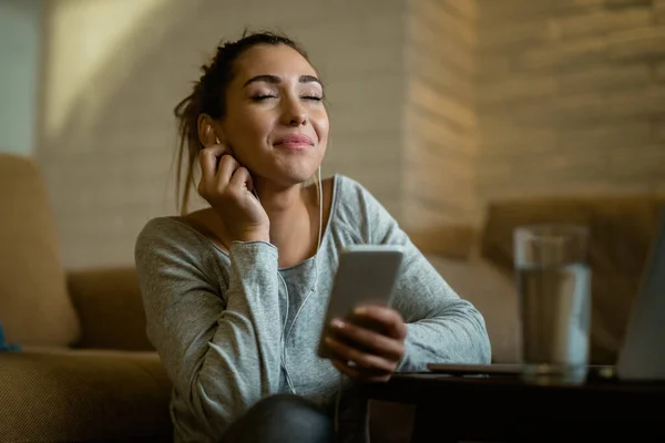 Smiling woman with eyes closed using smart phone and listening music in the evening at home.