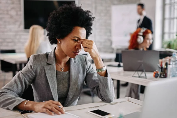 Tired black businesswoman reading an e-mail on a computer having a headache in the office. There are people in the background. 