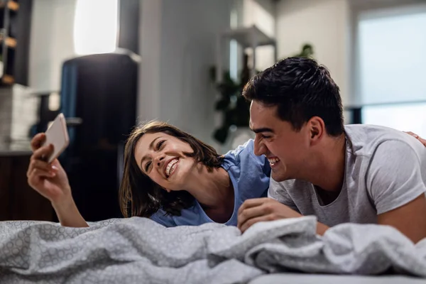Young happy woman using smart phone and taking selfie with her boyfriend while lying on the bed.
