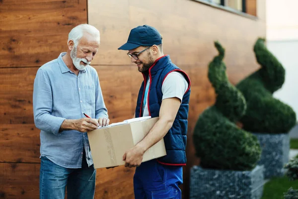 Happy senior man receiving packages and signing to postman for a home delivery.