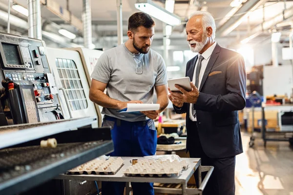 Smiling senior company manager showing something on digital tablet to manual worker in industrial facility. 