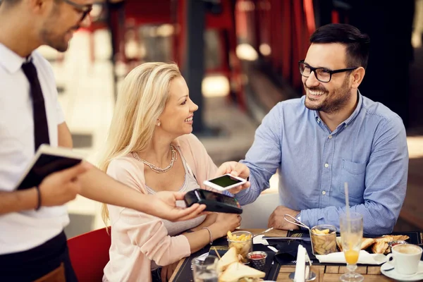 Happy couple making contactless payment with smart phone after lunch in a restaurant. 