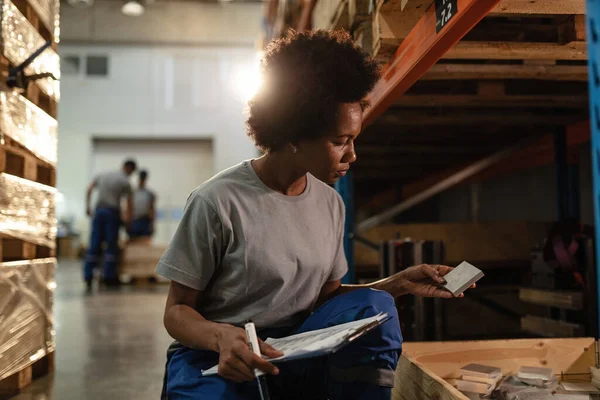African American warehouse worker going through check list while examining products in industrial storage compartment.