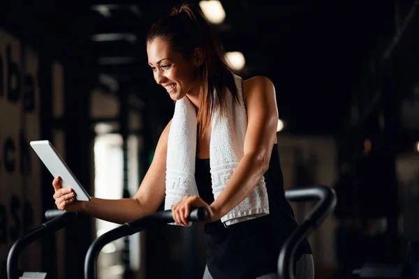 Happy athletic woman using digital tablet while working out on exercise bike in a gym.