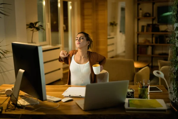 Young exhausted woman stretching while working late at night at home.