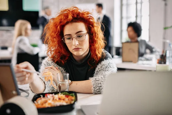 Young businesswoman feeling sad while having salad for lunch in the office. There are people in the background. 