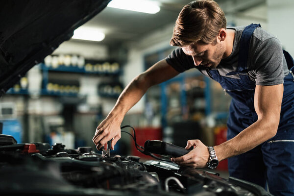 Young mechanic examining car battery with diagnostic tool while working in auto repair shop. 