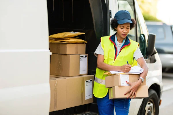 Black delivery woman filling shipment list while standing with packages next to a mini van.