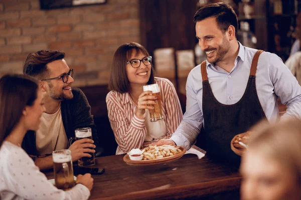 Happy waiter serving food to group of friends while they are drinking beer in a pub.