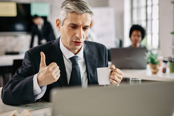 Satisfied businessman using laptop and showing thumbs up while reading good news. 