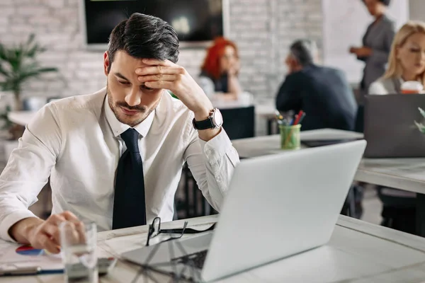 Pensive entrepreneur reading business reports while working in the office. There are people in the background. 