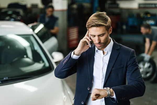 Young businessman checking the time on wristwatch while talking on mobile phone at auto repair shop.