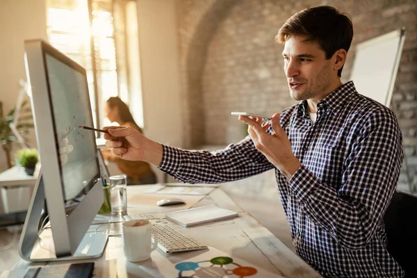 Happy businessman sending voice message on mobile phone while analyzing business progress on desktop PC.