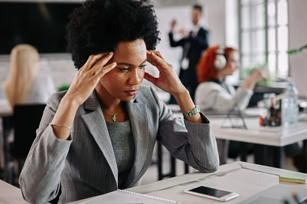 Pensive black businesswoman holding her head in pain while having some problems at work. There are people in the background.