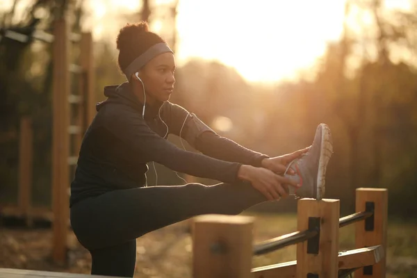Black female athlete stretching her leg while exercising at outdoor gym at sunset.