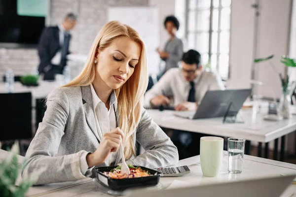 Mid adult businesswoman having lunch break at work and eating vegetable salad at her desk.