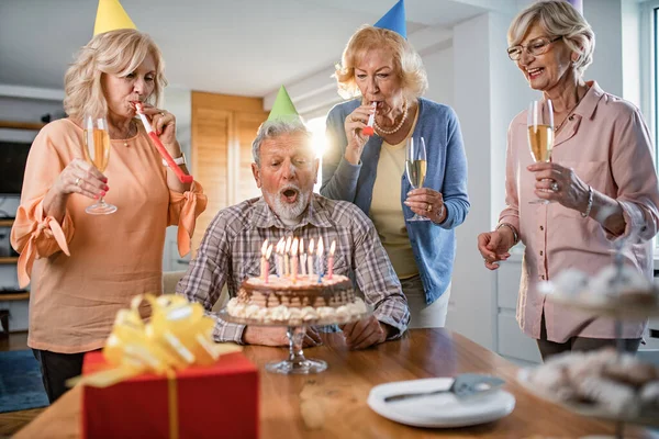 Mature birthday man blowing candles on the cake while celebrating birthday with his friends at home.