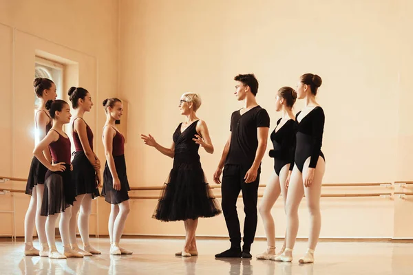 Mature ballet teacher talking to a group of dancers in ballet studio.