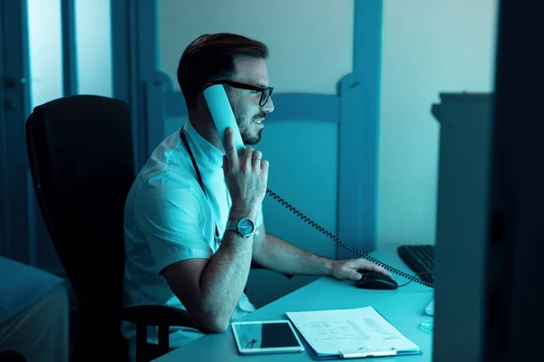 Male doctor working on a computer and communicating over the phone in the hospital.