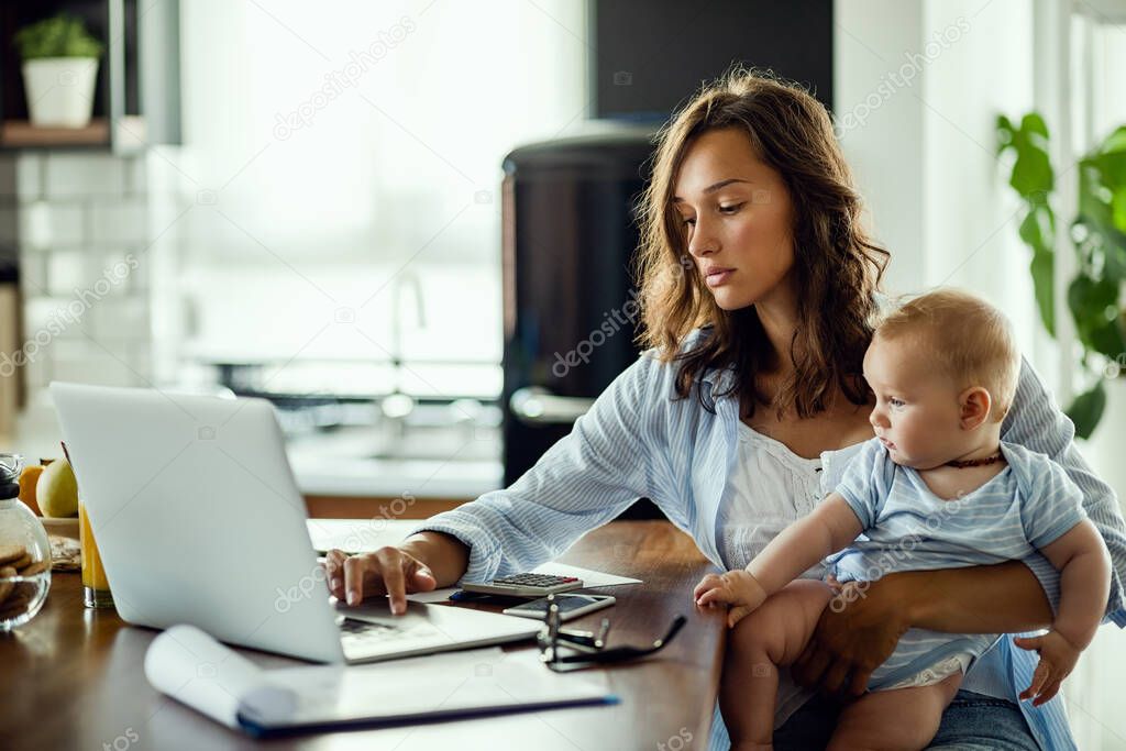 Young mother working on laptop while being at home with her baby son.